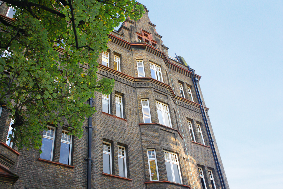 An old brown brick building with multiple windows, with a tree in front of the building, and a blue sky behind it.